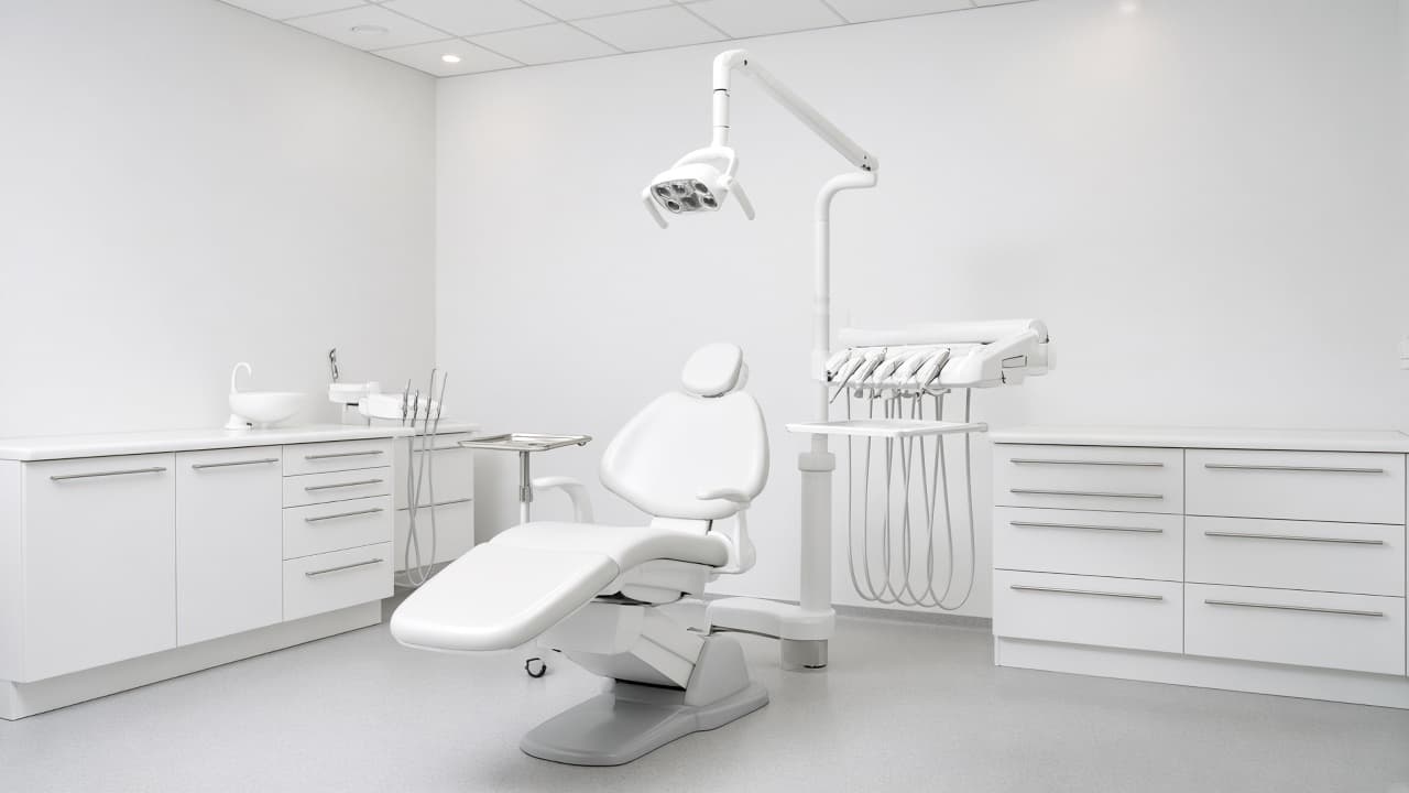 A high-key photograph of an empty all-white operatory with an upright dental chair and an overhead light lowered above it.