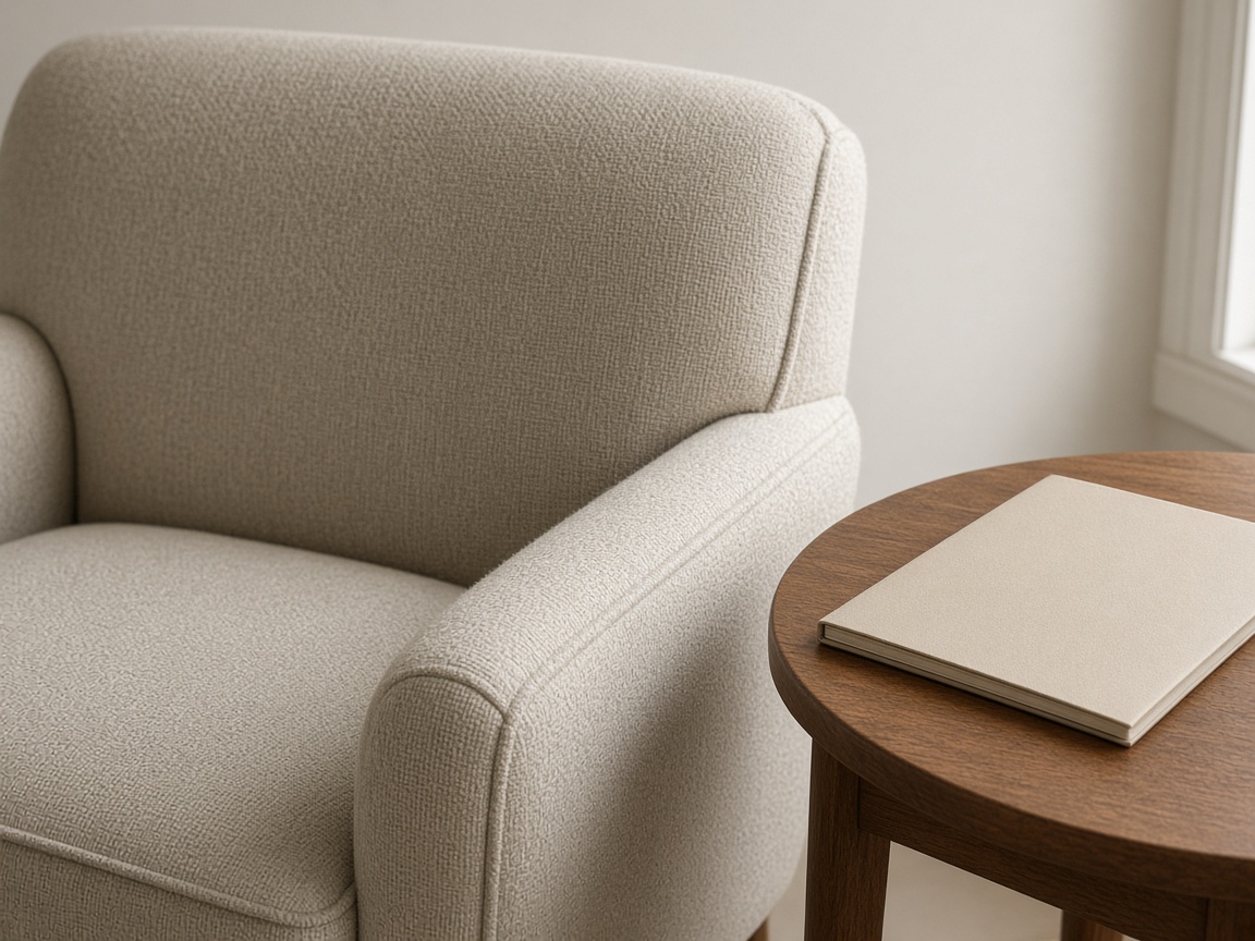 Close view of an oatmeal armchair arm and a closed cream folder on a round wood side table.